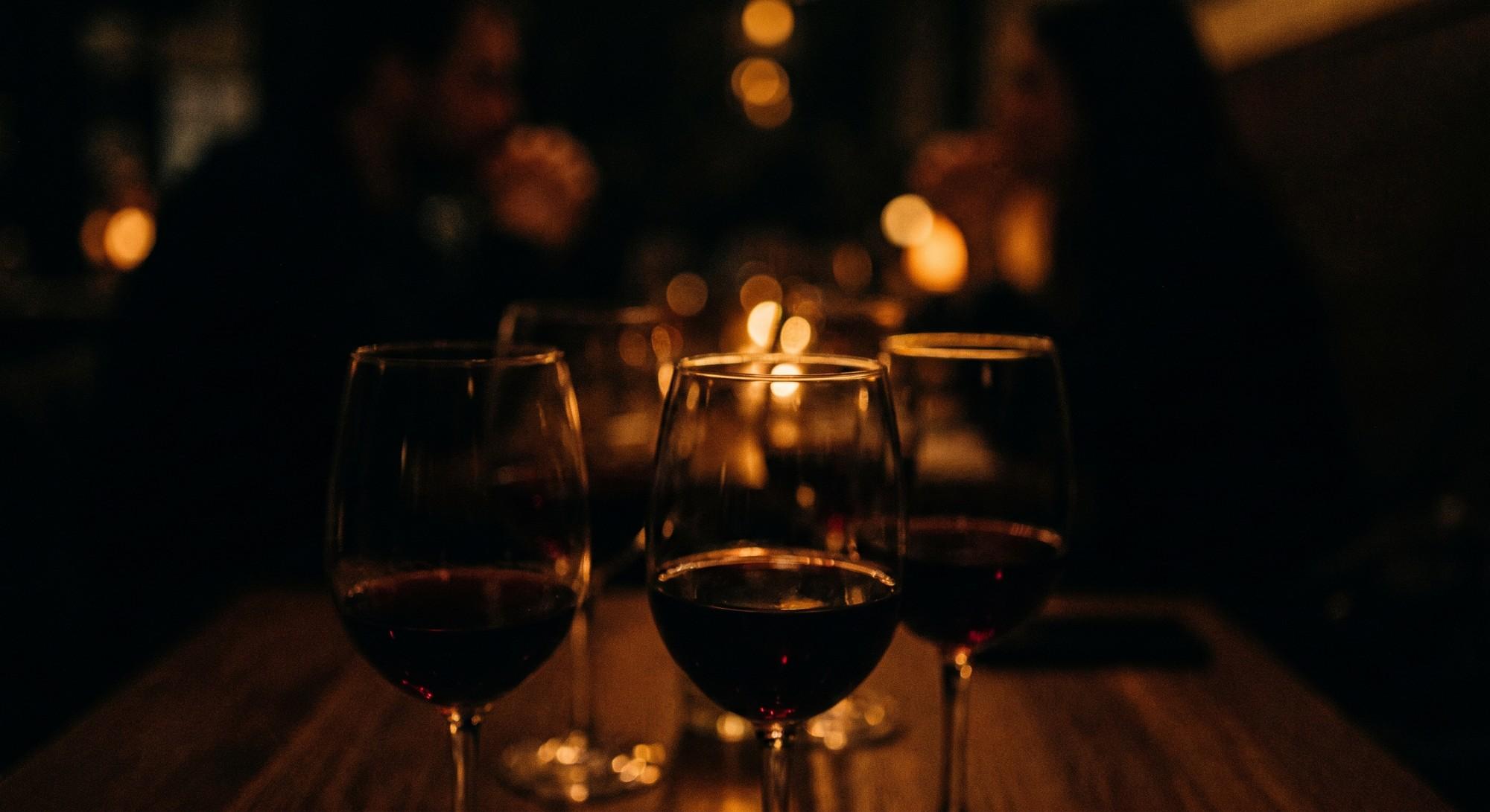 Three glasses of red wine on a wooden table in candlelight, guests blurred behind.